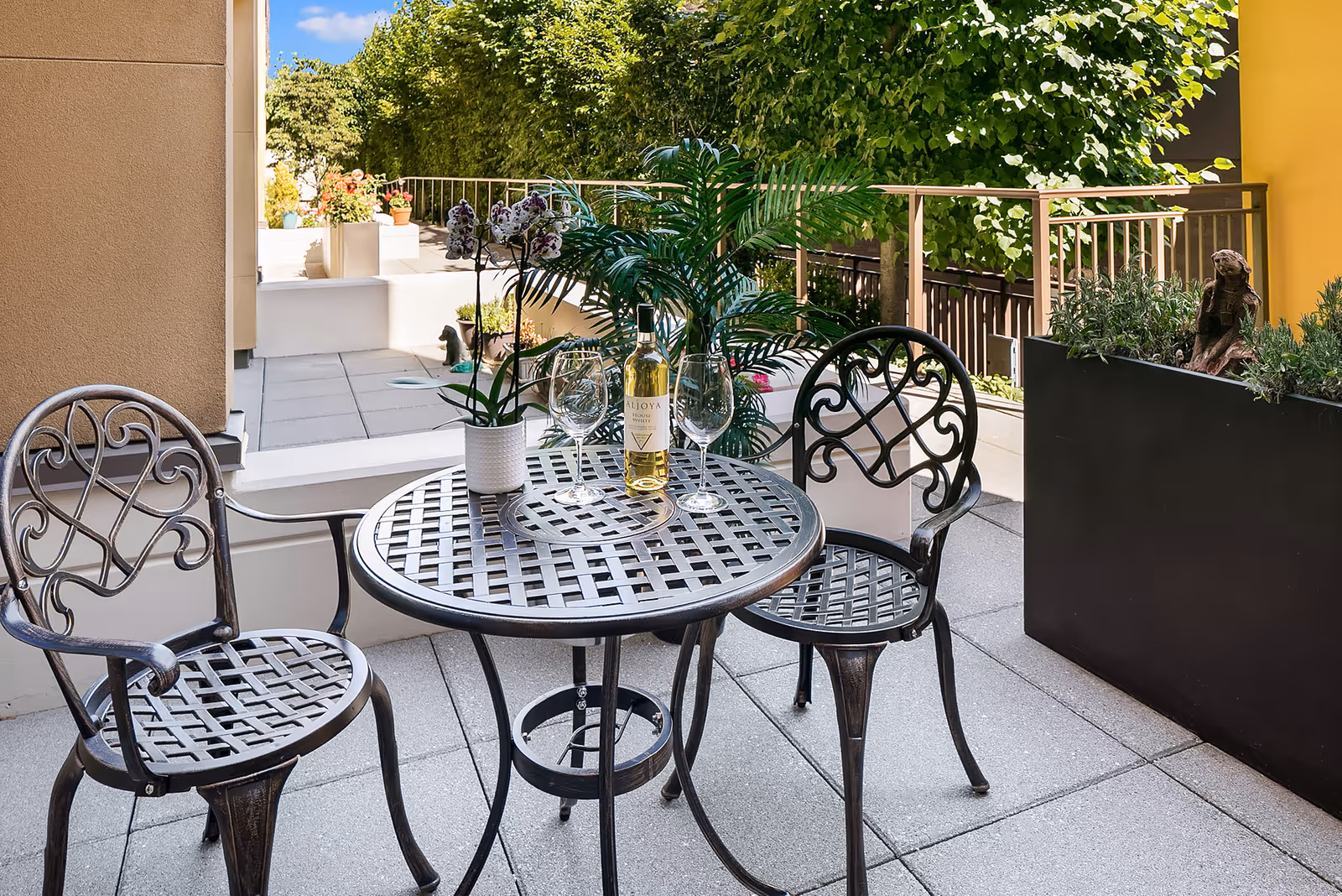 Outdoor patio area with a round metal table and two matching chairs. On the table, there is a bottle of white wine labeled 'Aljoya' and two empty wine glasses, along with a potted orchid plant. The patio is surrounded by greenery and potted plants, with a railing and a yellow wall on one side.