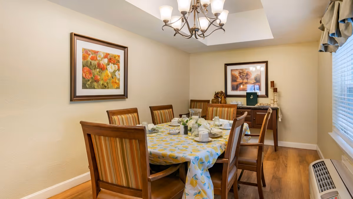 A dining room with a rectangular wooden table covered with a yellow and blue patterned tablecloth. Six wooden chairs with striped cushions surround the table, which is set with white plates, cups, and napkins. The room has light-colored walls, two framed paintings, a chandelier overhead, a window with blinds and a valance, and a wooden sideboard with decorative items.