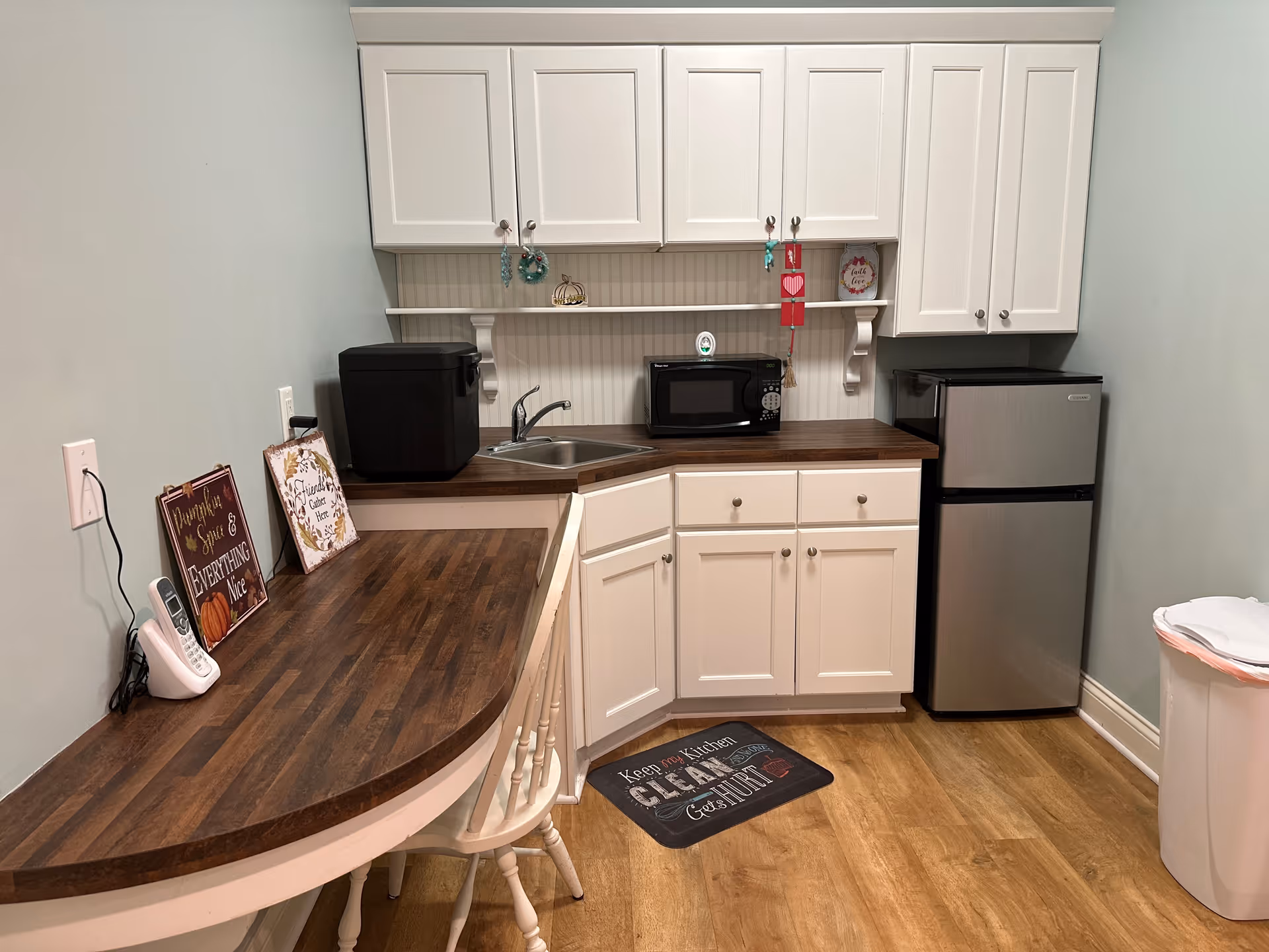 Compact kitchenette with white cabinets, wood countertops, a sink, microwave, mini fridge, and a small breakfast bar with chairs.