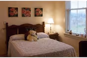 A cozy bedroom with a wooden headboard bed covered in a white quilt. A teddy bear is placed on the bed. There is a bedside table with a lamp and a window with blinds letting in natural light. Three floral paintings hang on the wall above the bed.