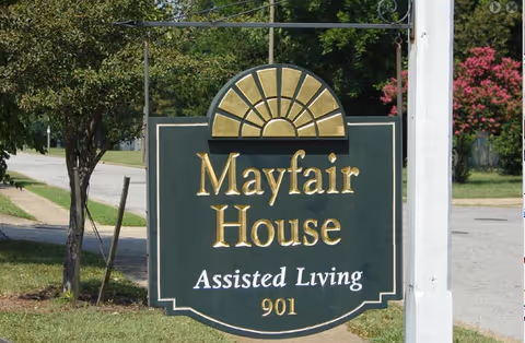 Green wooden sign that reads 'Mayfair House Assisted Living 901' hanging outdoors near a street.