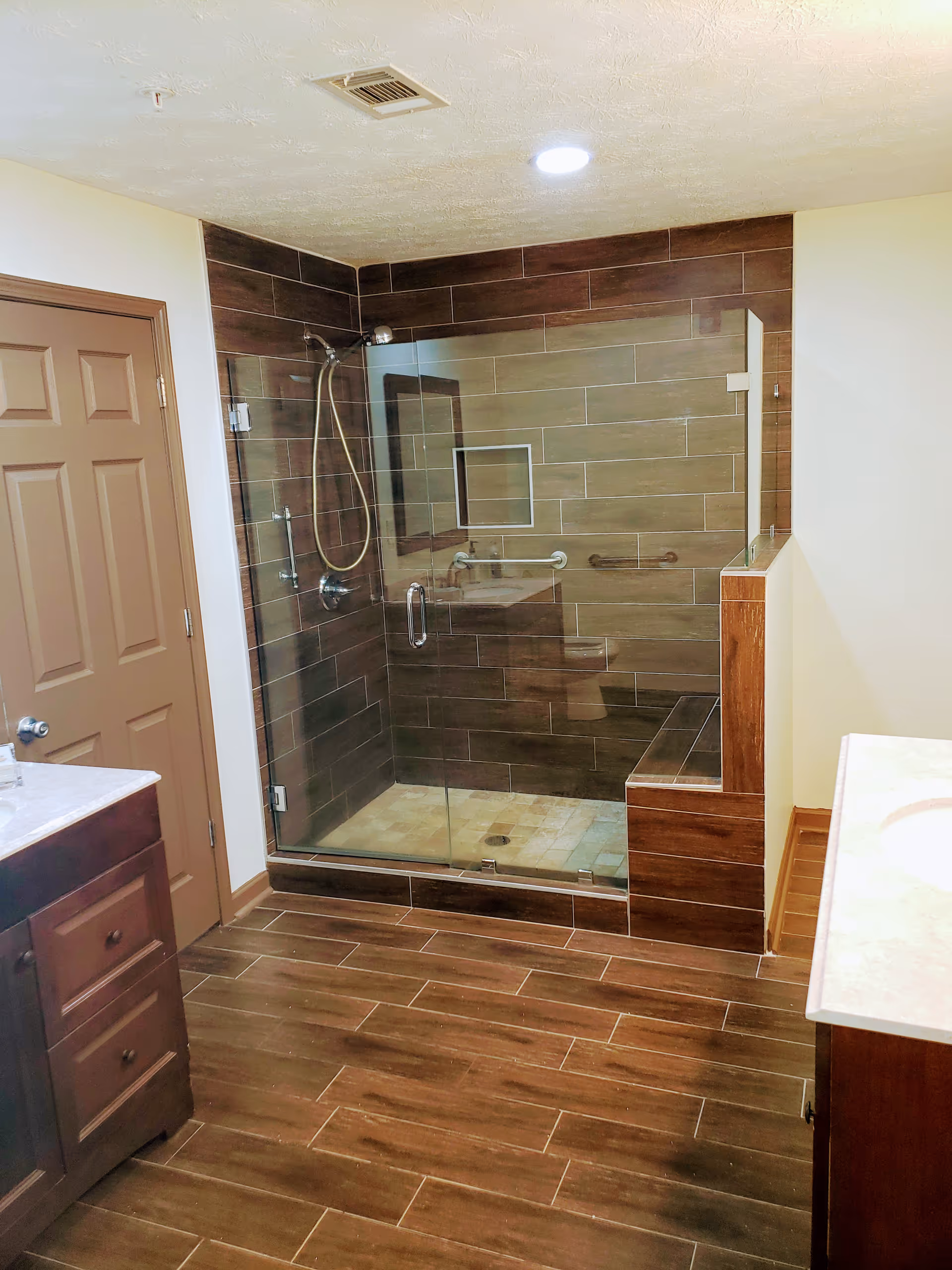 Glass-enclosed tiled walk-in shower with bench and handheld showerhead in a bathroom with wood-tone floor and vanities.