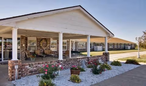Exterior view of a single-story building with a covered porch supported by white columns. The porch has seating and a decorative wreath on the brick wall. In front of the porch is a landscaped area with white gravel, green plants, and pink flowers. Another similar building is visible in the background under a clear sky.