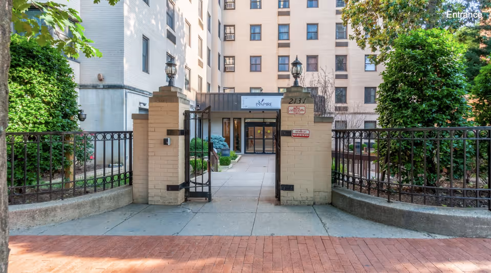 Entrance gate to Inspire Rehabilitation & Health Center with beige brick pillars and black metal fencing, leading to the main building entrance surrounded by greenery and trees.