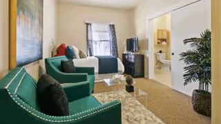 A cozy living area in an assisted living facility featuring two green armchairs with black pillows, a glass coffee table on a beige rug, a bed with white and green bedding in the background, a TV on a stand, and a potted plant near a sliding door that opens to a bathroom.