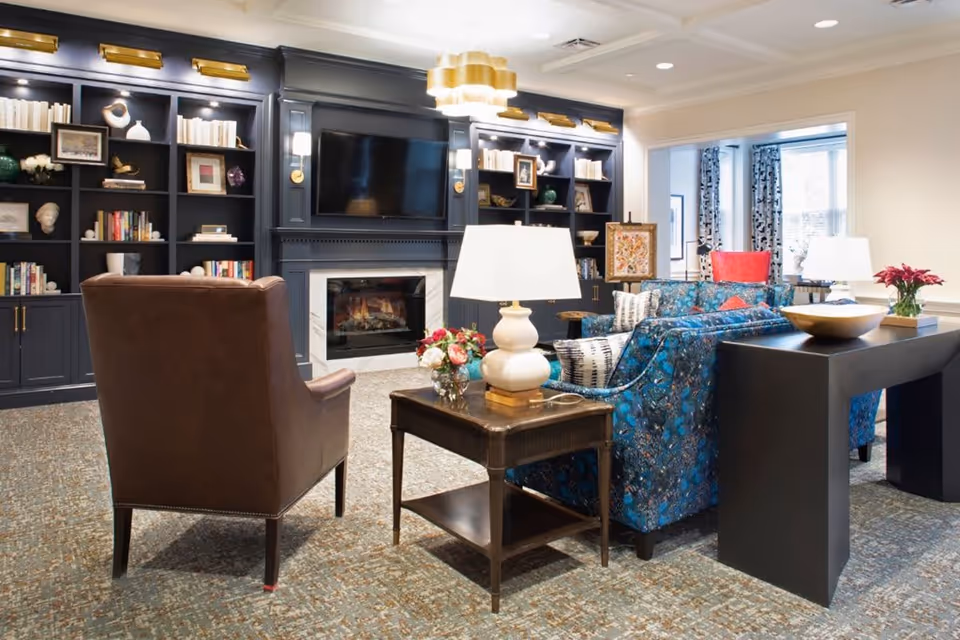 A cozy living room with a dark blue built-in bookshelf filled with books and decorative items, a flat-screen TV mounted above a fireplace, a brown leather armchair, a blue patterned sofa, a wooden side table with a white lamp and flowers, and a black console table with a bowl and flowers. The room has carpeted flooring and large windows with patterned curtains.