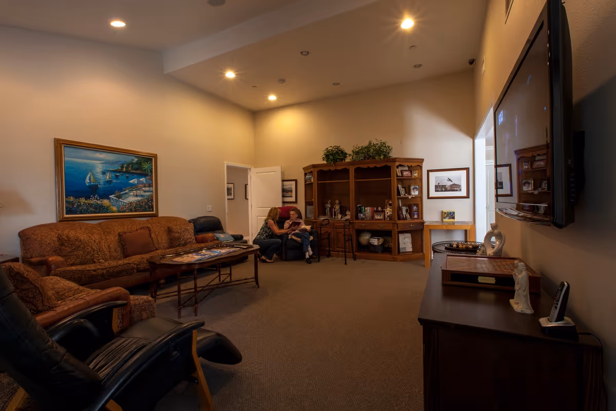 A cozy communal living room with sofas, armchairs, a wall-mounted TV, wooden shelving and two people seated near the back.