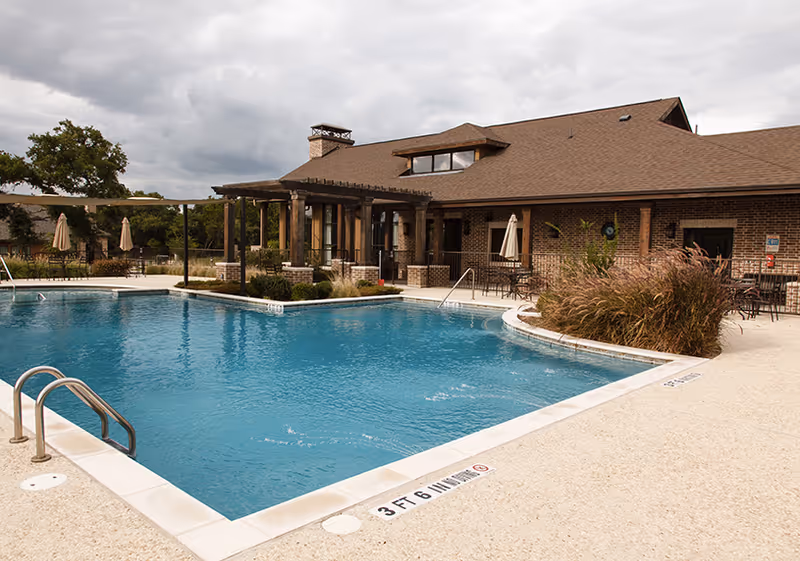 Outdoor swimming pool with clear blue water next to a brick building with a brown roof. The pool area has metal handrails, patio tables with umbrellas, and landscaping with bushes and trees under a cloudy sky.