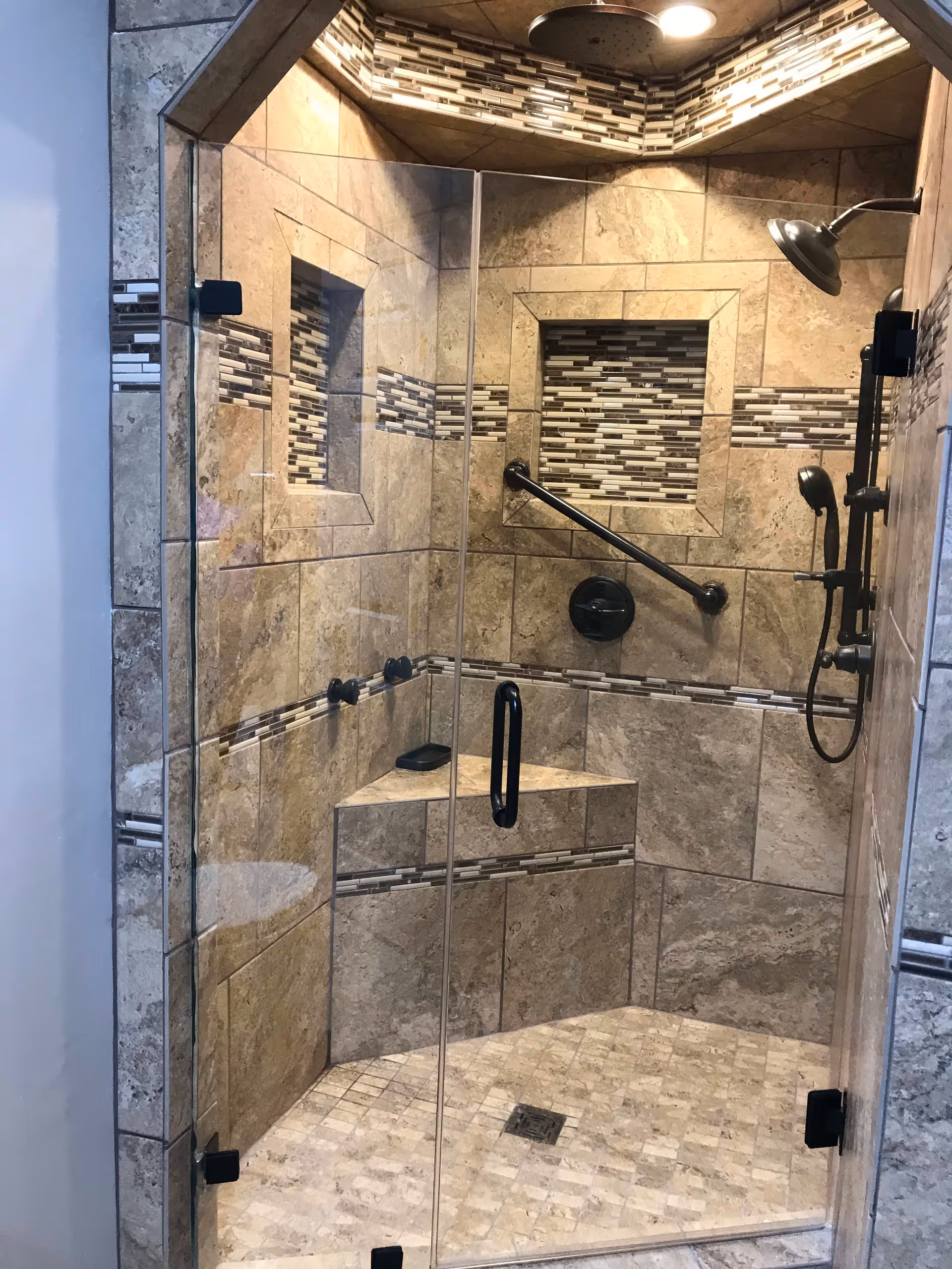 A modern tiled shower with a glass door, built-in bench, and decorative tile accents. The shower features a rainfall showerhead, a handheld showerhead, a grab bar, and two recessed niches for storage.