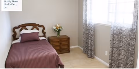 A simple bedroom with a single bed featuring a wooden headboard and a purple bedspread. Next to the bed is a wooden nightstand with a vase of flowers. The room has beige walls, a tiled floor, and a window with patterned curtains allowing natural light to enter.