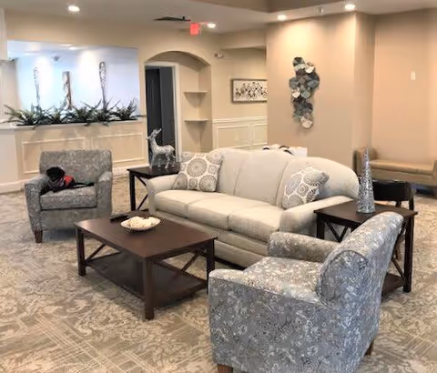 A cozy living room area with a beige sofa, two patterned armchairs, a dark wooden coffee table, and side tables. Decorative items including a small sculpture and a bowl are on the tables. The room has neutral-colored walls, carpeted floor, and some wall art and plants in the background.