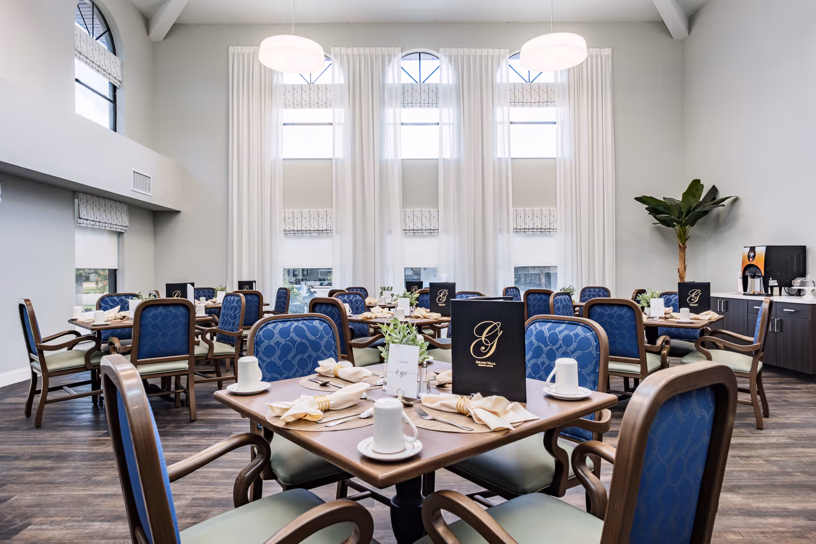 A bright and spacious dining room with multiple wooden tables set with white cups, plates, napkins, and silverware. Each table has blue cushioned chairs with wooden frames. Large windows with white curtains allow natural light to fill the room. There are menus on the tables labeled 'Grand Villa of Englewood' and a beverage station with a coffee machine and cups on the right side. A tall green plant is placed near the beverage station.