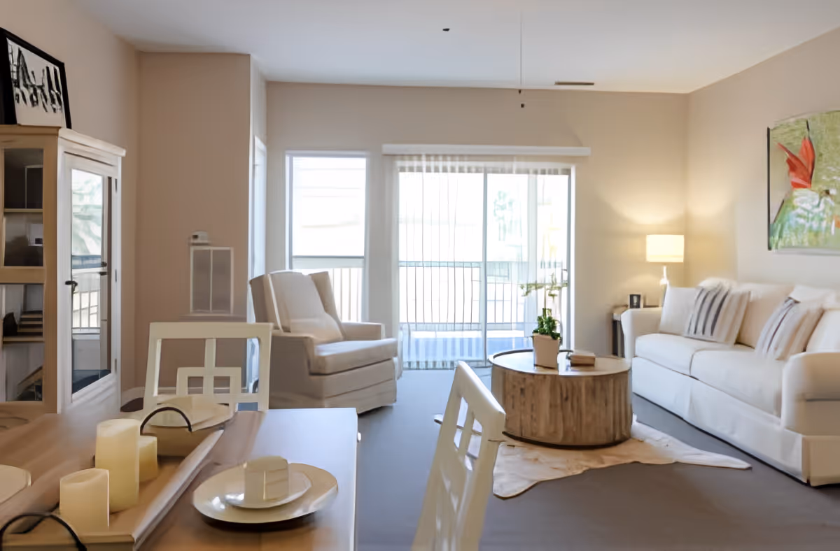 Bright neutral-toned living room and dining area with a sofa, armchair, round wooden coffee table, dining table with candles, and sliding glass doors to a balcony.