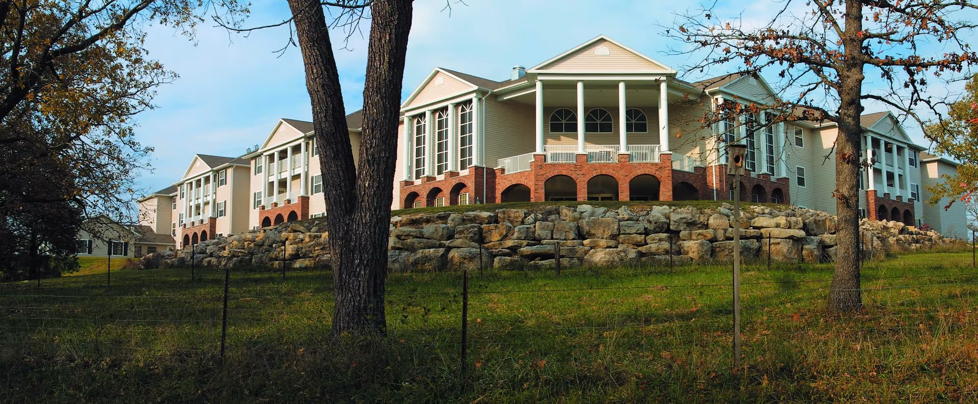 Front exterior of a multi-story retirement community building on a rocky hill with trees and grassy foreground.