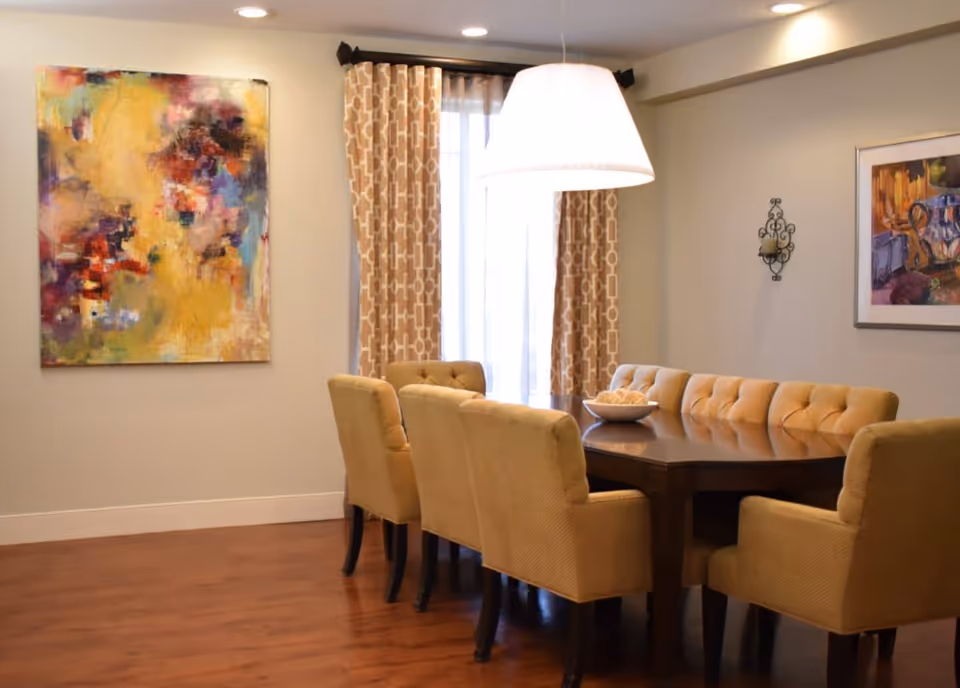 A dining room with a dark wooden table surrounded by eight beige upholstered chairs. The room has wooden flooring, patterned beige curtains covering a window, and two colorful abstract paintings on the walls. A large white pendant light hangs above the table.
