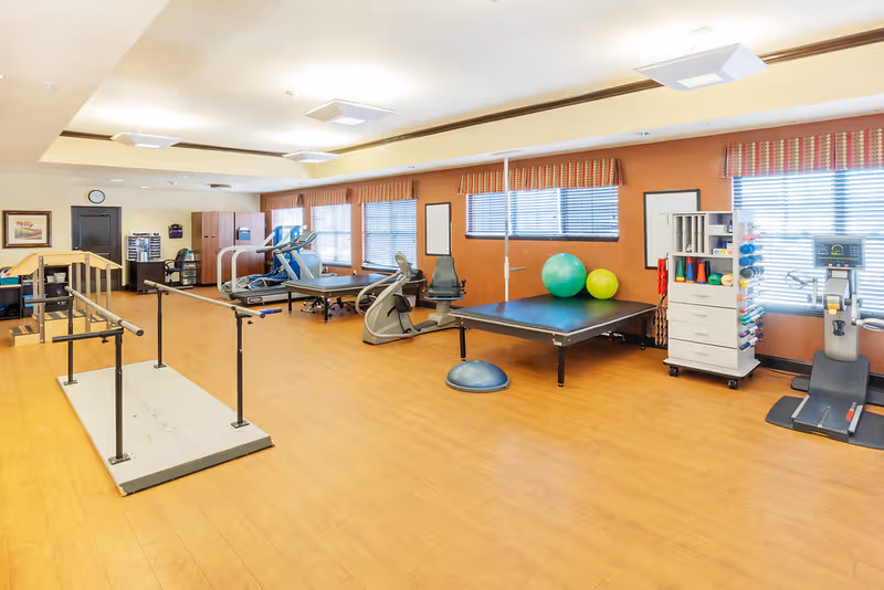 A spacious rehabilitation room with wooden flooring and orange walls, equipped with parallel bars for walking exercises, treadmills, stationary bikes, therapy tables, exercise balls, and various physical therapy equipment. Large windows with blinds allow natural light to fill the room.