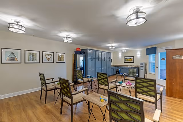 A well-lit room with wooden flooring featuring six chairs arranged in a semi-circle around small tables. The chairs have green patterned backs and beige cushions. On the tables are small colorful dumbbells. The back wall has a row of blue lockers and a counter with cabinets. The walls are decorated with framed artwork and there are ceiling lights providing illumination.