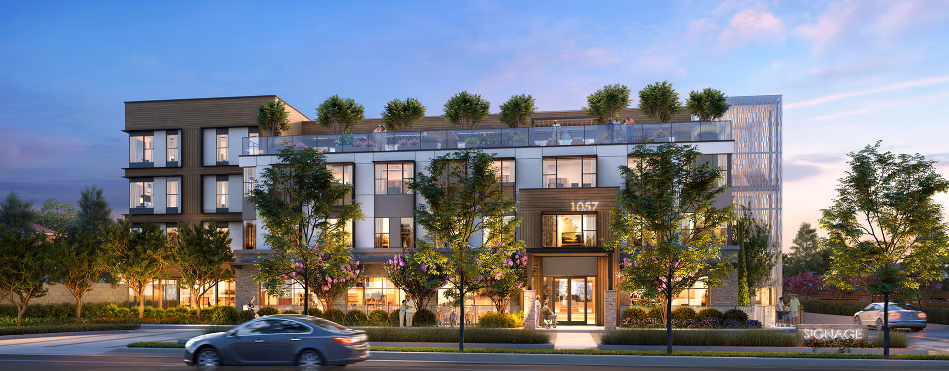 Exterior view of a modern three-story assisted living and memory care facility at dusk with large windows, trees, and landscaped greenery. People are visible on the rooftop terrace and near the entrance. Cars are driving on the street in front of the building.