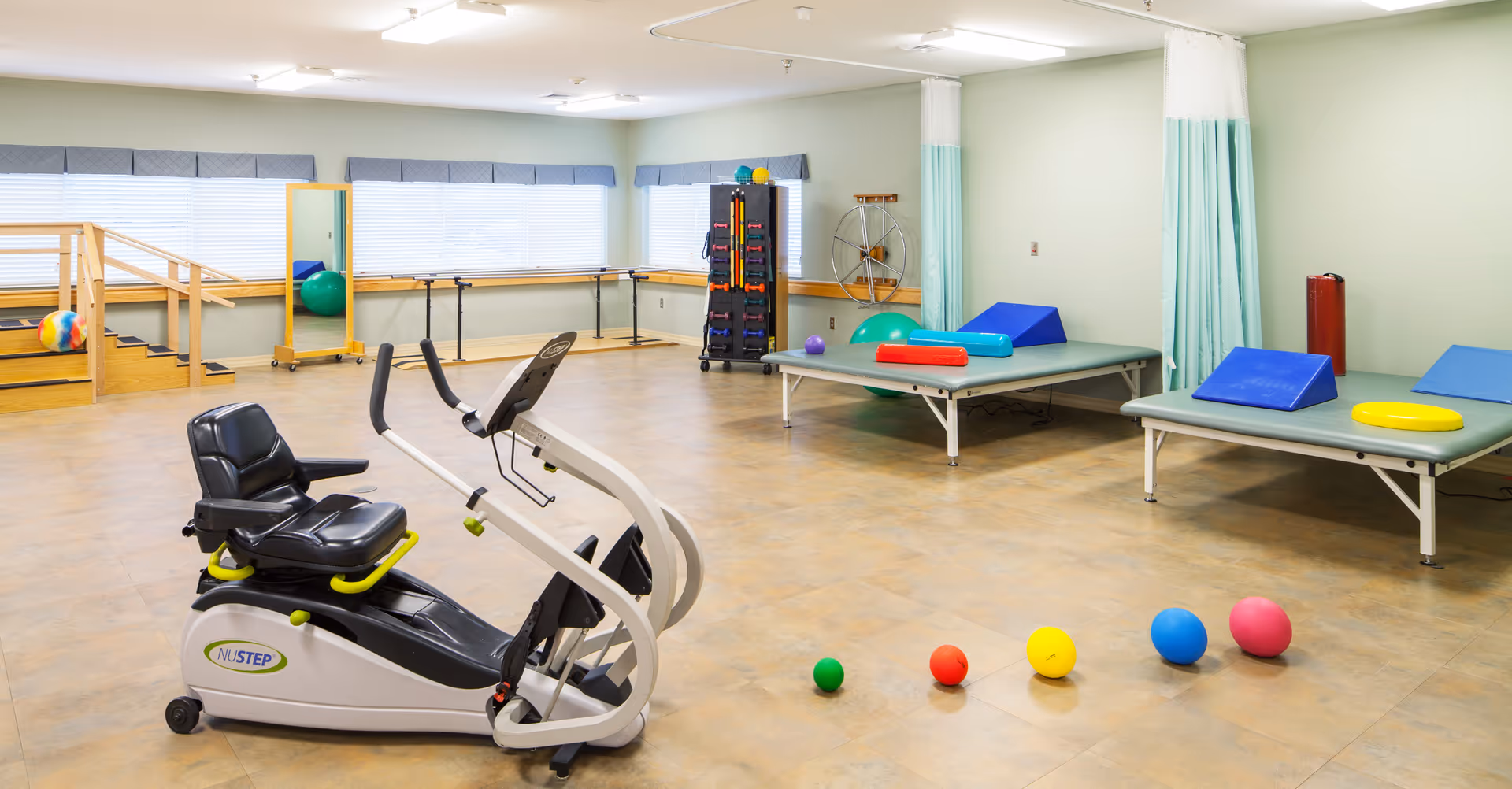 A spacious physical therapy or rehabilitation room with exercise equipment including a recumbent exercise bike, therapy tables with colorful cushions, various therapy balls, a wooden ramp with stairs, a large mirror, and handrails along the walls. The room has large windows with blinds and light green walls.