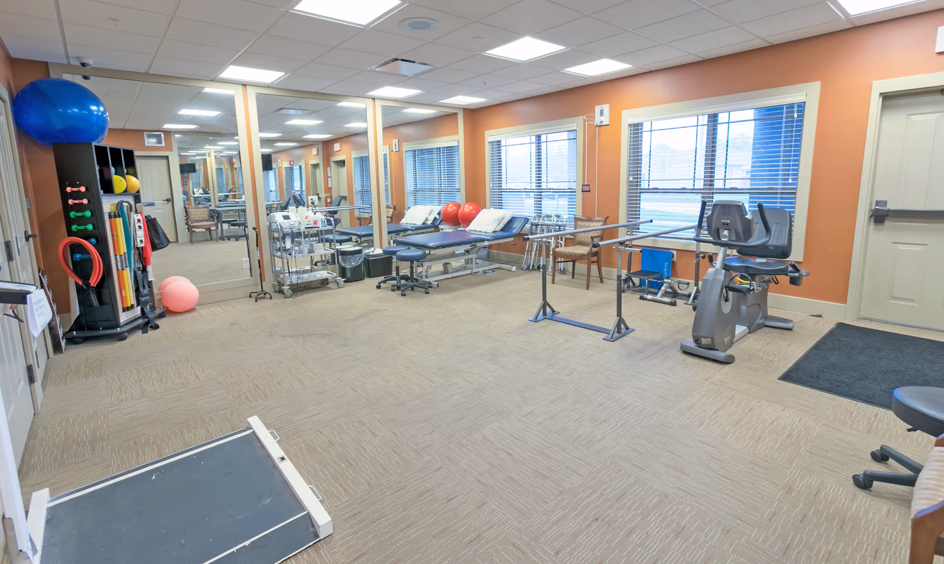 A physical therapy or rehabilitation room with exercise equipment including a recumbent bike, parallel bars, therapy tables with pillows, exercise balls, and various therapy tools organized on shelves. The room has large windows with blinds, mirrors on one wall, and beige carpeting.