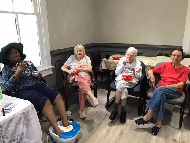 Four elderly women sitting in chairs in a room with gray walls and wood flooring. One woman on the left is wearing a green hat and using a foot spa, while the others are eating from red bowls. There is a window on the left side letting in natural light and a table with a tablecloth and some items in the background.