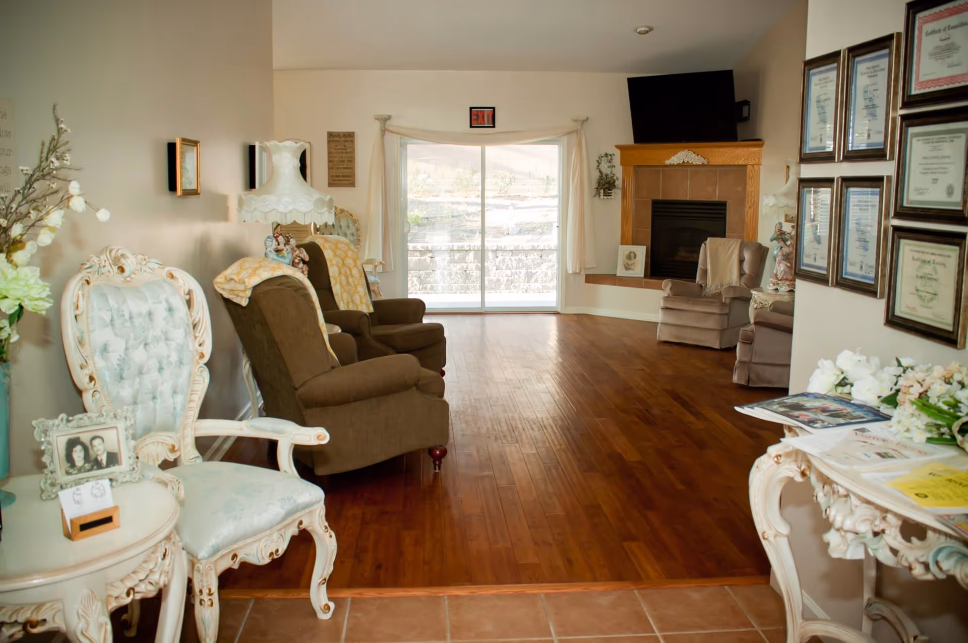 A cozy living room with wooden flooring, featuring several upholstered armchairs and a vintage white chair with a small table holding a framed photo. There is a fireplace with a TV mounted above it, certificates framed on the wall, and a sliding glass door leading outside. The room is decorated with flowers and soft lighting.
