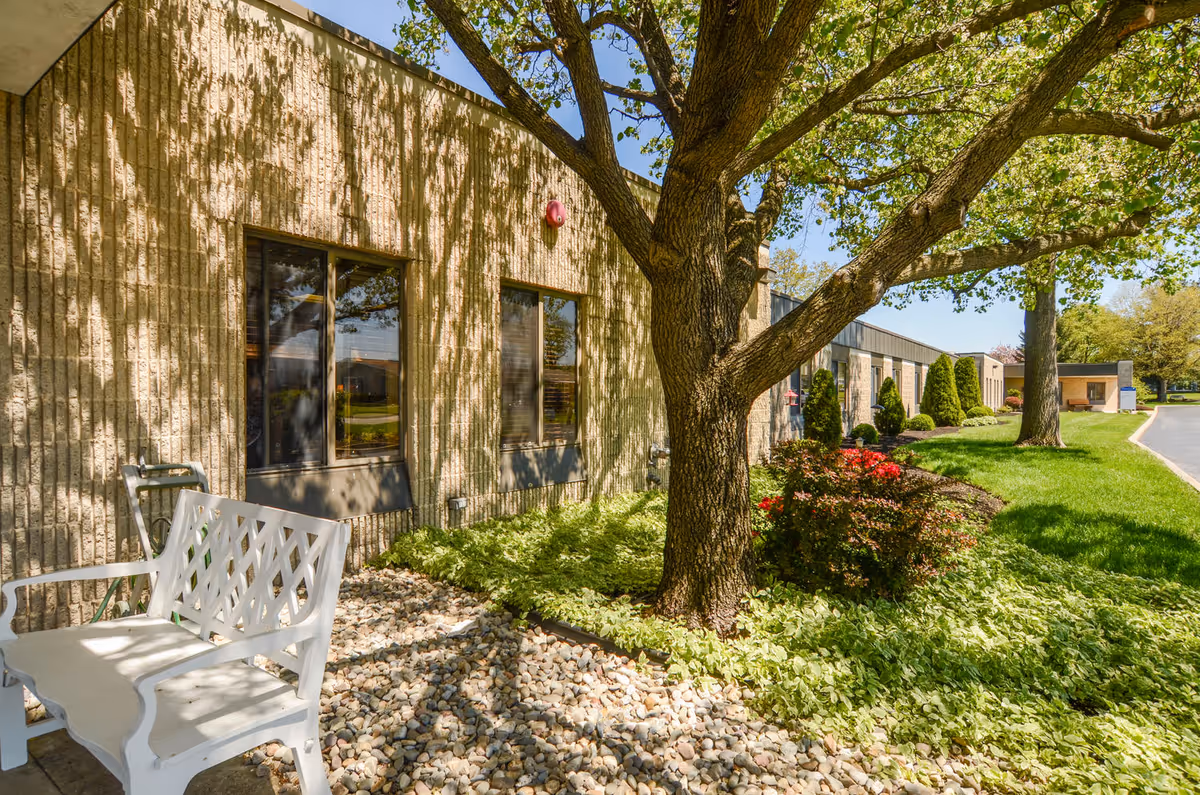 Exterior view of a single-story senior care building with a large tree, landscaped rocks and shrubs, and a white bench.