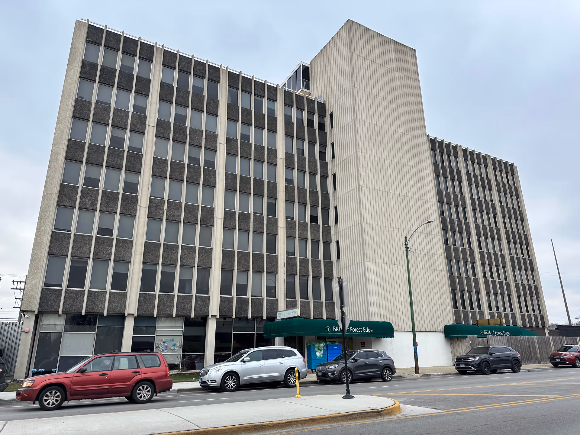 Exterior view of a multi-story building with a gray and beige facade, featuring many windows. The building has green awnings above the entrances with the text 'BRIA of Forest Edge'. Several cars are parked along the street in front of the building under a cloudy sky.