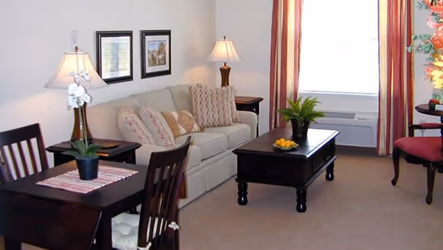 A cozy living room with a beige sofa adorned with patterned cushions, a dark wooden coffee table with a potted plant and a bowl of lemons, a small dining table with two chairs and a potted plant, two table lamps on side tables, framed artwork on the wall, and a window with striped curtains letting in natural light.