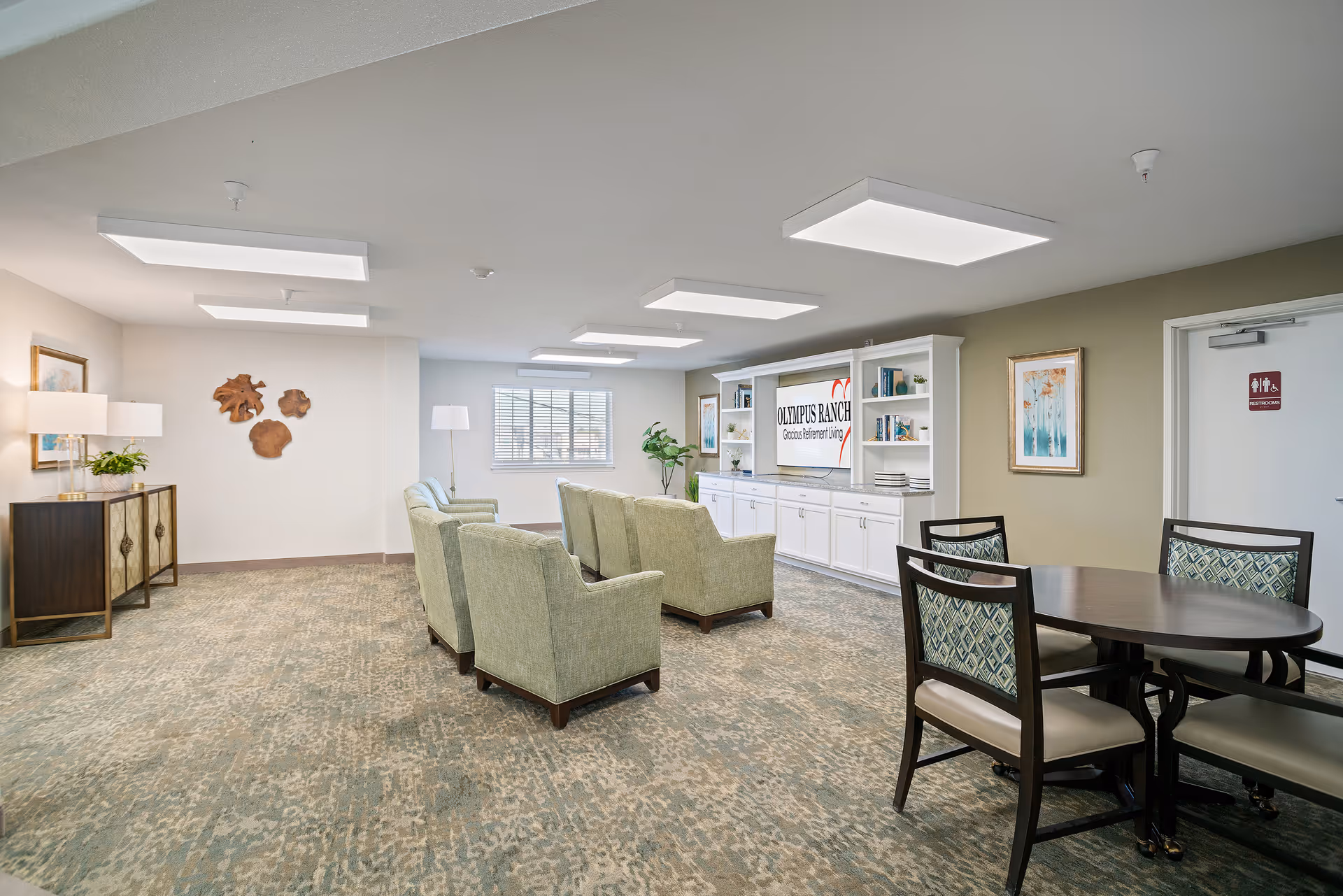 Carpeted communal lounge with a cluster of green armchairs, a round table with chairs, built-in white shelving displaying an "Olympus Ranch" sign, and wall decor.
