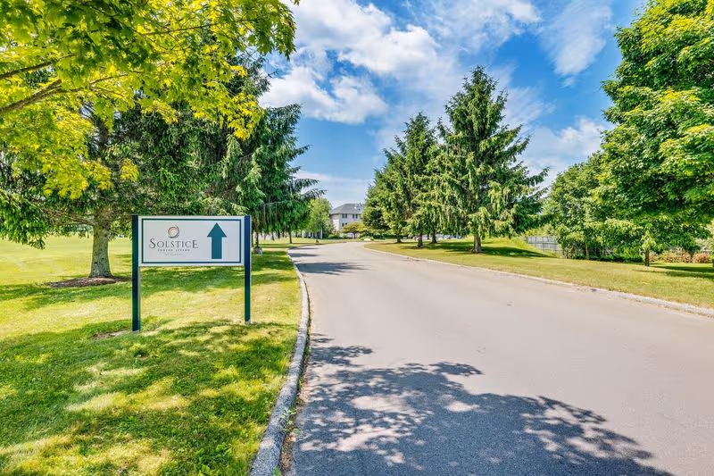 A paved driveway lined with green grass and tall trees on both sides leading to a building in the distance under a partly cloudy blue sky. A sign on the left side of the driveway reads 'Solstice Senior Living' with an arrow pointing straight ahead.