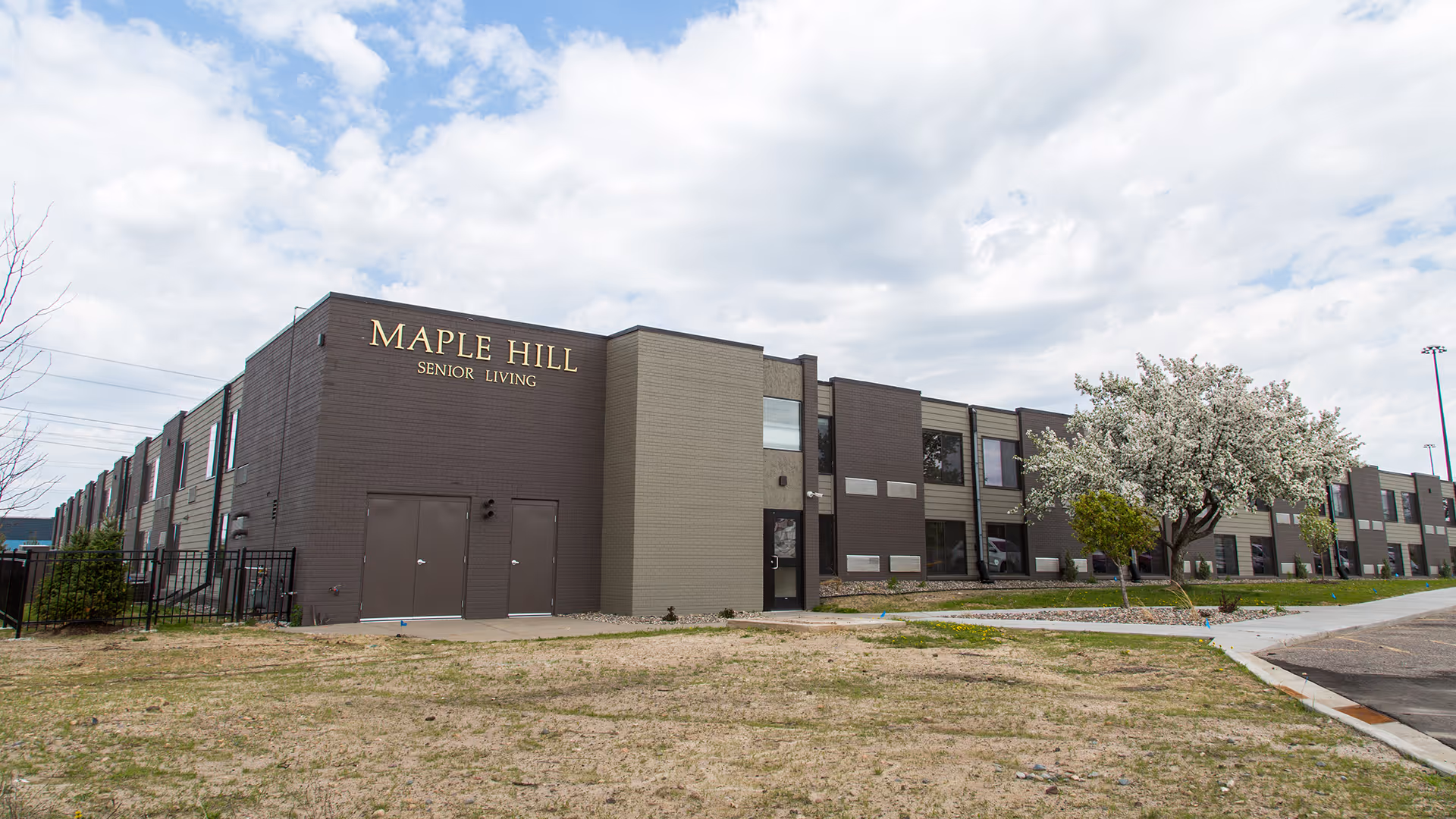 Exterior view of Maple Hill Senior Living facility, a two-story building with brown and beige walls, several windows, and a flowering tree near the entrance under a partly cloudy sky.