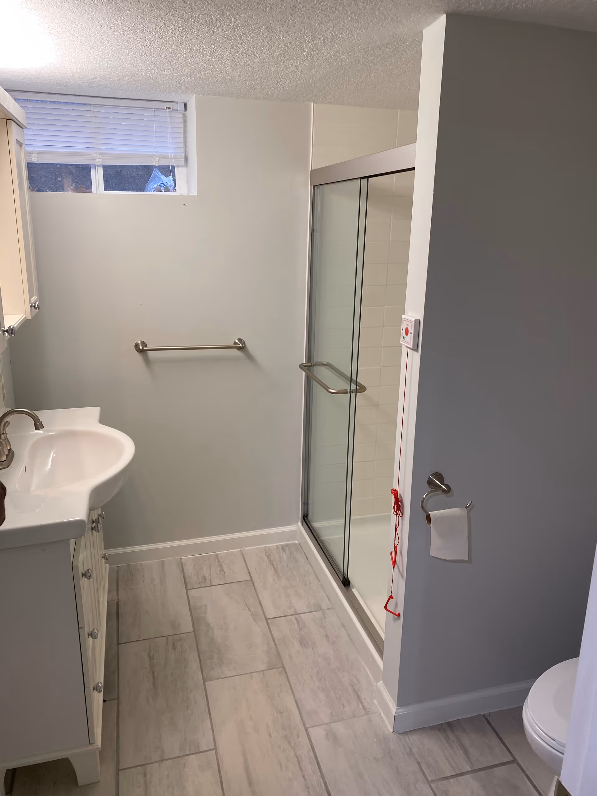Bright small bathroom with a vanity sink, glass-enclosed shower, towel bar, toilet, and a red emergency pull cord.