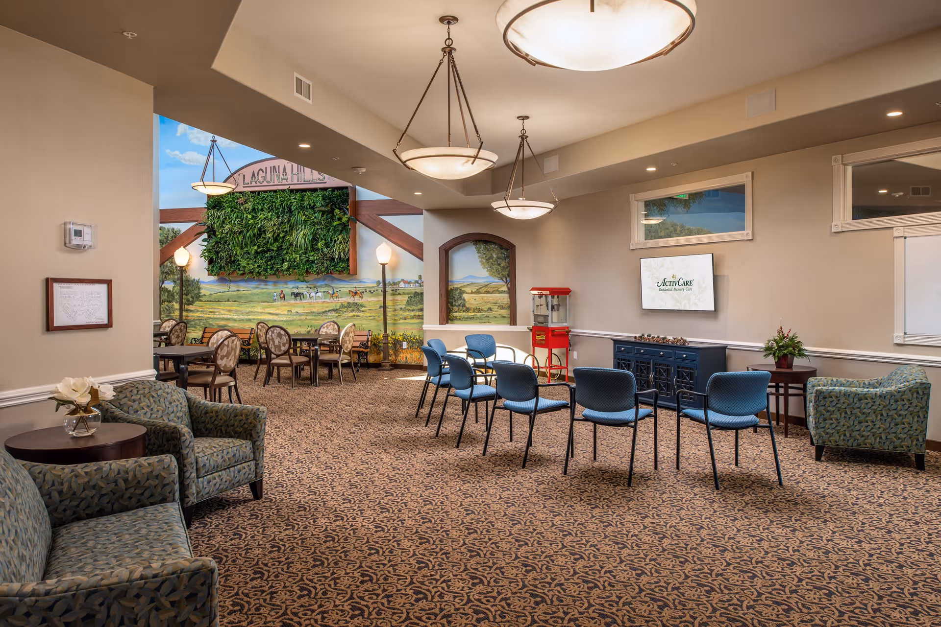 A spacious, well-lit common area in a senior living facility with patterned carpet, multiple seating arrangements including blue chairs in rows facing a TV screen displaying 'ActivCare Residential Memory Care', armchairs with side tables, and a red popcorn machine. The back wall features a mural of a countryside scene with horses and people, and a green vertical garden with the text 'LAGUNA HILLS' above it.