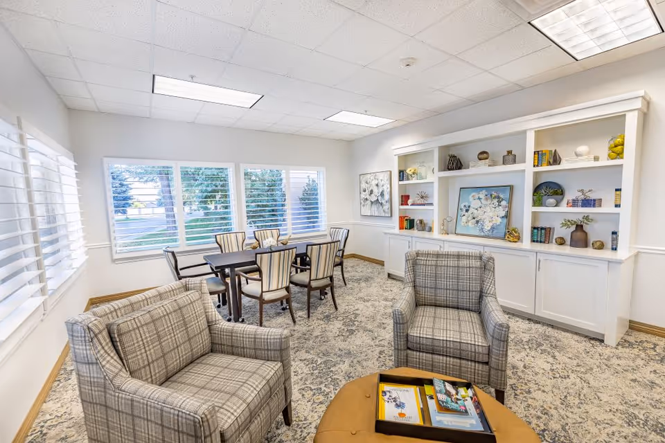 A bright and cozy common area in a retirement residence featuring two plaid armchairs and a small round table with magazines in the foreground. In the background, there is a dining table with six chairs near three large windows with white blinds. A white built-in bookshelf with decorative items and artwork is against the wall.
