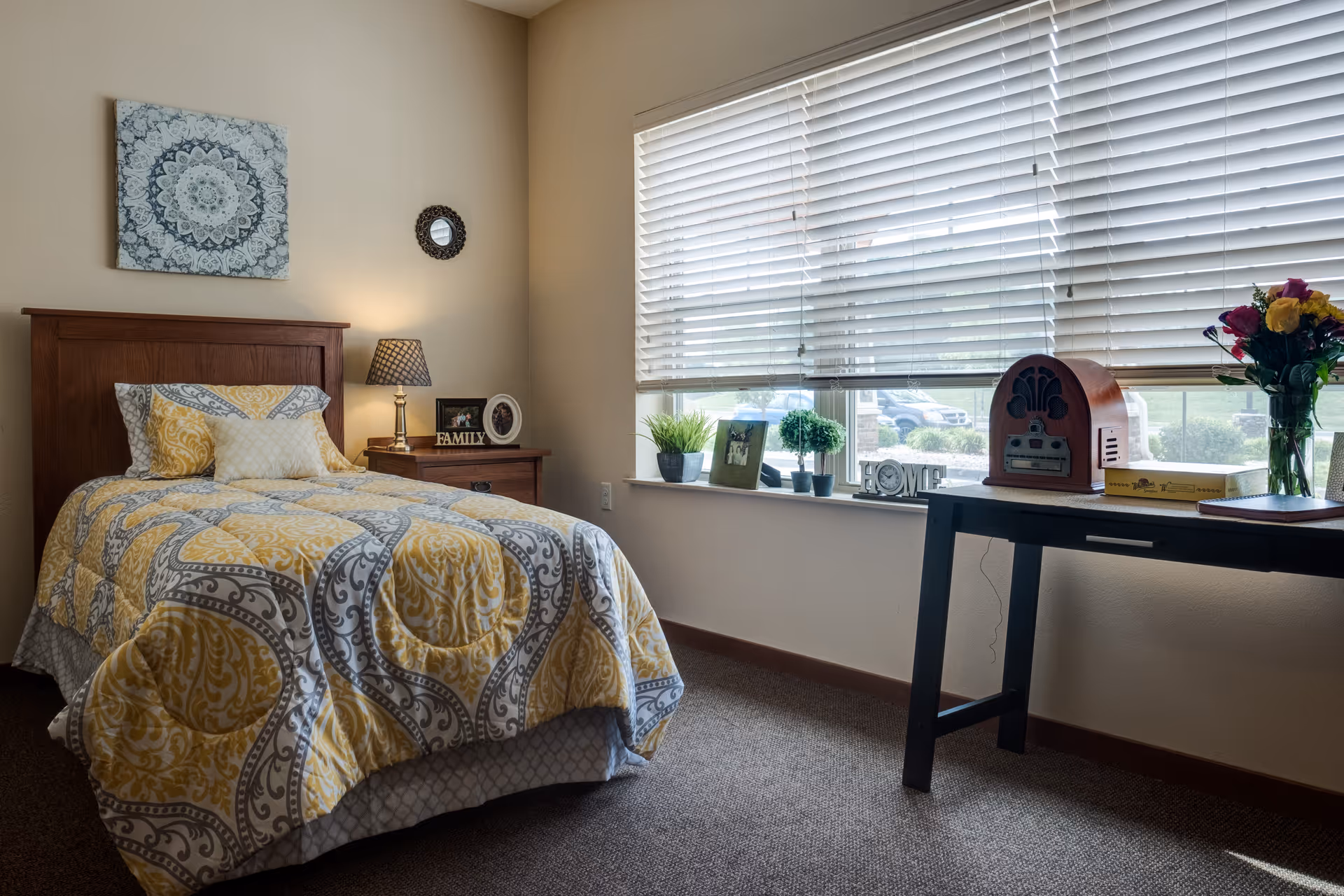 A cozy bedroom with a single bed covered in a yellow and gray patterned comforter. Next to the bed is a wooden nightstand with a lamp, framed photos, and a decorative 'FAMILY' sign. A large window with white blinds lets in natural light, and a black table beneath the window holds a vintage radio, a box, a vase of colorful flowers, and small potted plants. The walls are decorated with a blue and white mandala artwork and a small round mirror.