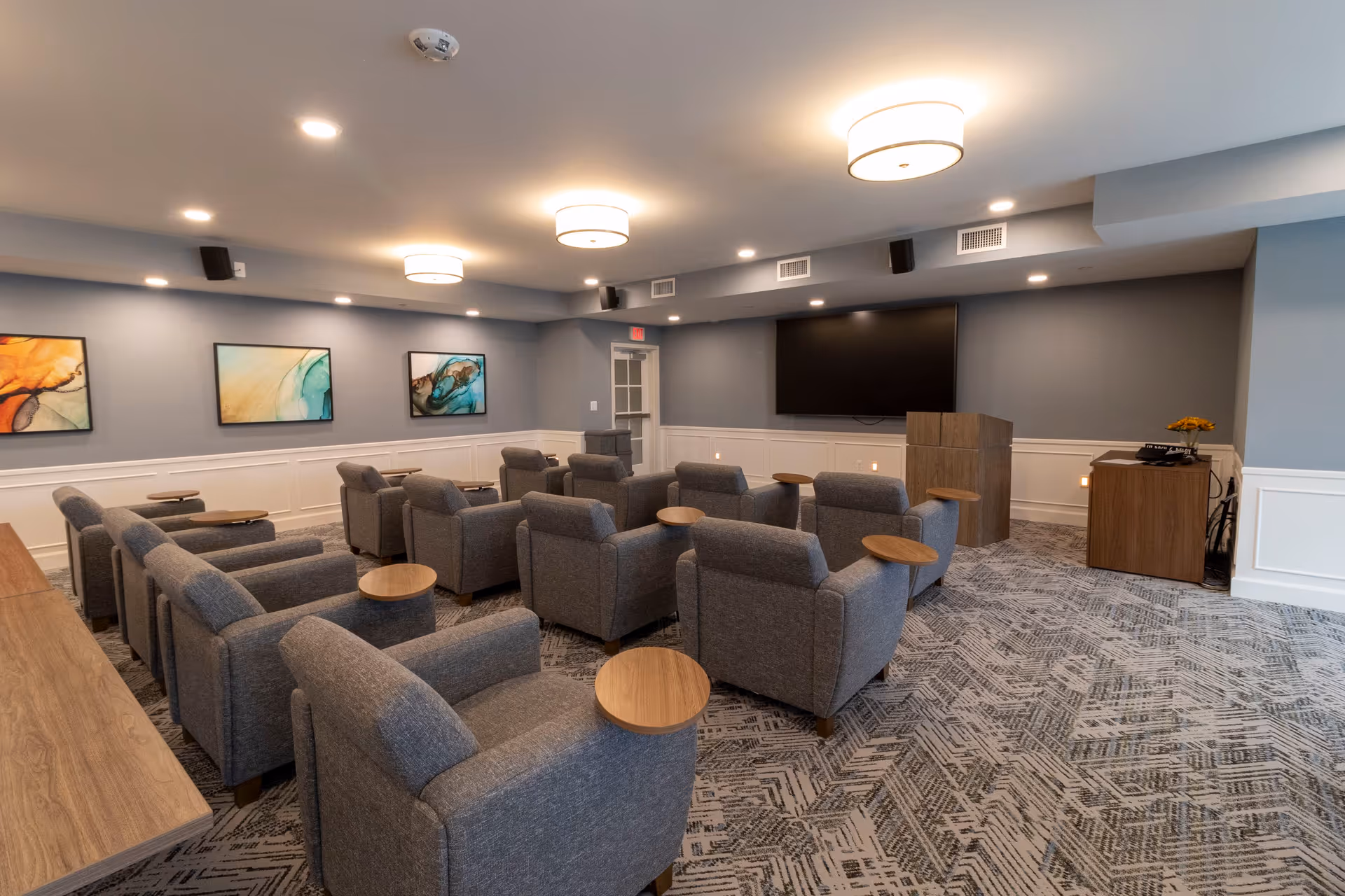 A cozy media room with rows of gray armchairs each with small round side tables facing a large wall-mounted TV and podium.