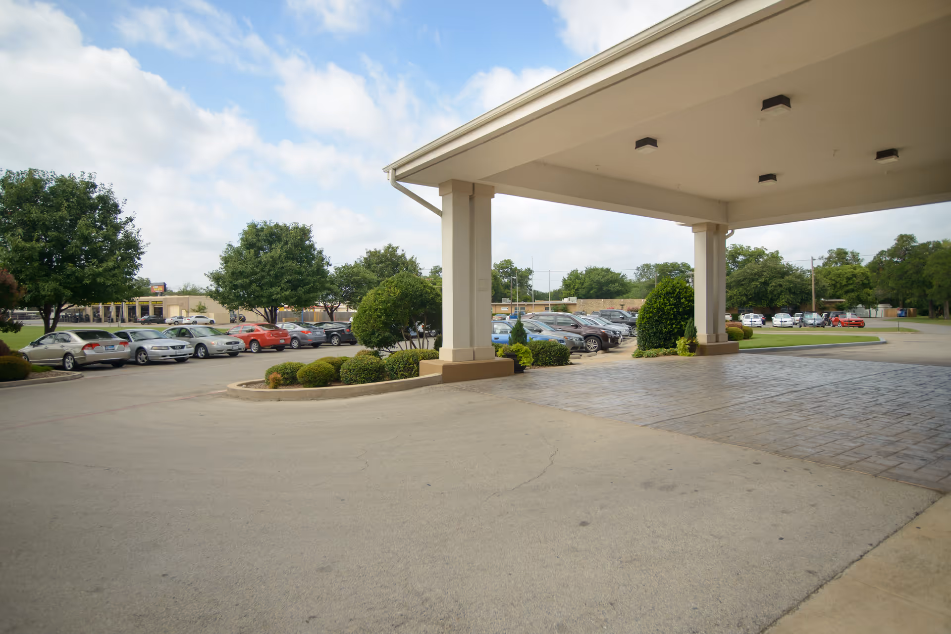 Covered entrance area of a facility with a paved driveway and parking lot filled with cars. There are landscaped bushes and trees around the parking area under a partly cloudy sky.