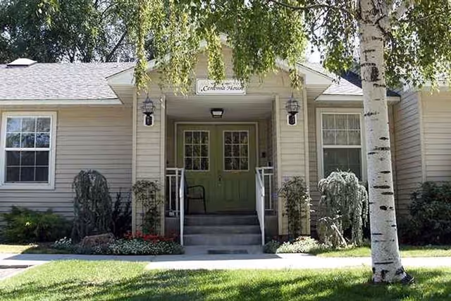 Front exterior view of Cenoma House, a single-story building with beige siding, green double doors, two windows on either side, a small porch with steps and railings, outdoor lantern-style lights, and surrounding greenery including trees and shrubs.