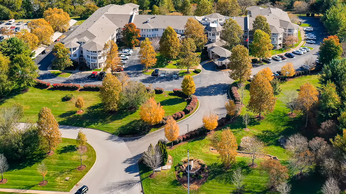 Aerial view of Manor at Steeplechase, a senior living community, showing multiple connected buildings surrounded by landscaped grounds with trees displaying autumn colors, parking lots with cars, and curved driveways.
