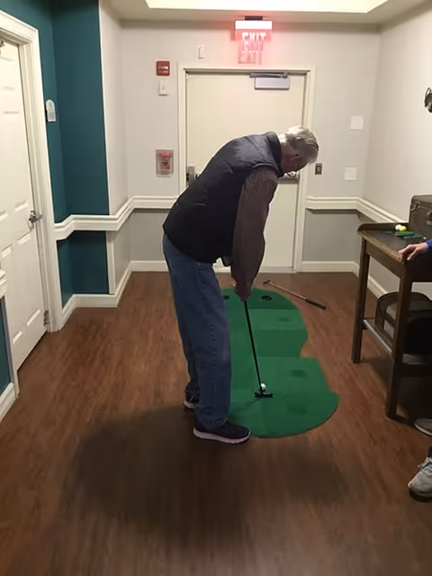 An elderly man playing indoor mini golf on a green putting mat in a hallway with wood flooring and light-colored walls. There is a table with a stool to the right and an exit door in the background.