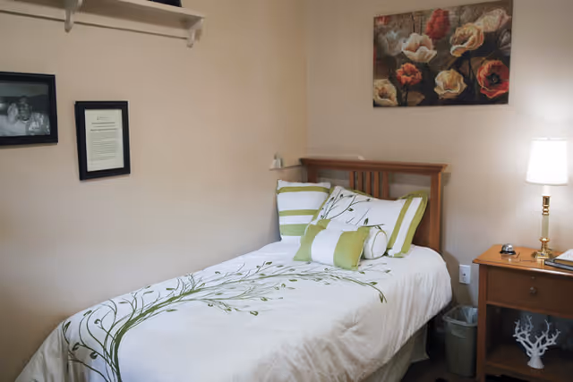 Small single bedroom with a twin bed dressed in white and green floral bedding, a wooden nightstand with a lamp, and wall art above the headboard.