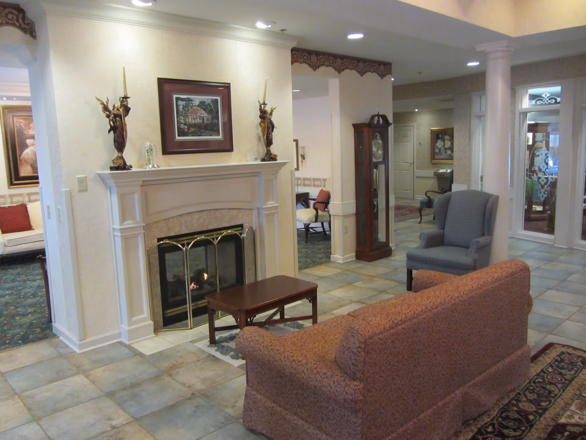 A cozy assisted living facility common area with a fireplace, a small wooden coffee table, a patterned sofa, and a blue armchair. The room has tiled flooring, decorative wall art, a grandfather clock, and columns near the entrance.