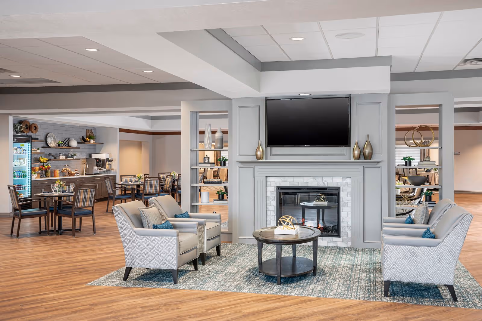 A modern senior living facility common area featuring a cozy seating arrangement with four upholstered armchairs around a round wooden coffee table on a patterned rug. Behind the seating area is a decorative fireplace with a mounted flat-screen TV above it and shelves with vases on either side. In the background, there is a dining area with tables and chairs, and a refreshment station with a refrigerator and coffee machine.
