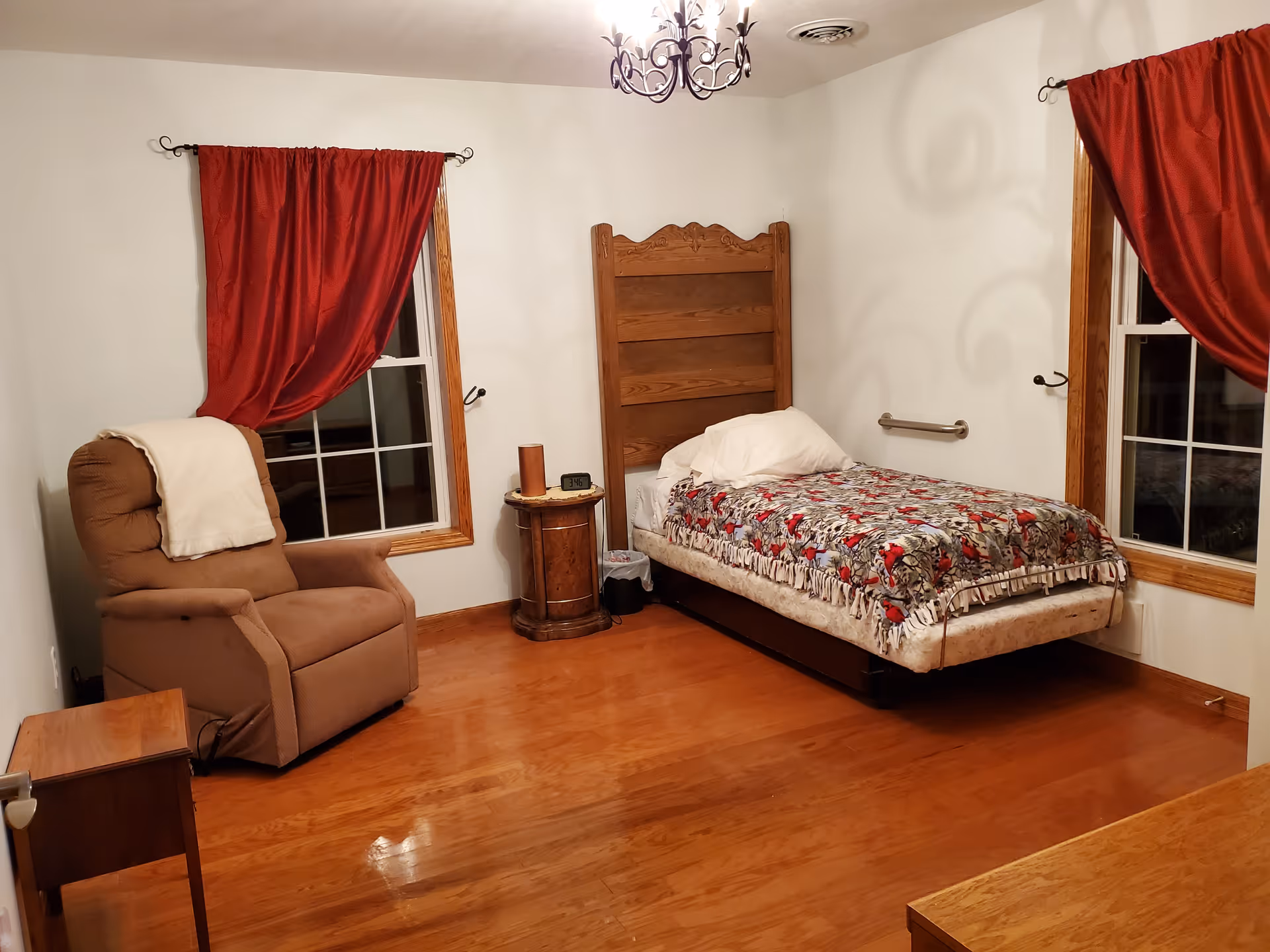 A simple bedroom with a single bed, wooden headboard, recliner chair, side table, hardwood floor, and red curtains over two windows.