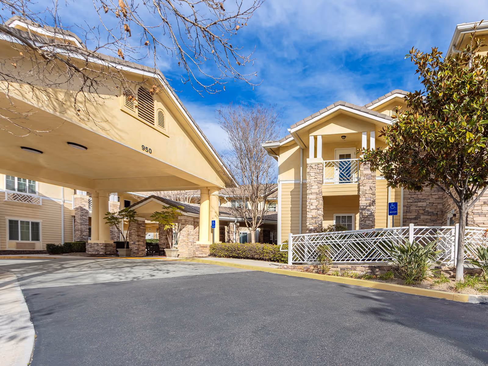 Exterior view of a senior living facility with beige buildings, stone accents, a covered entrance with the number 950, trees, and a clear blue sky.