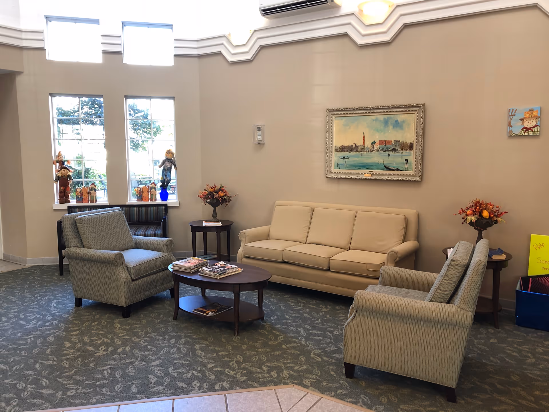 A cozy living room area with a beige sofa, two patterned armchairs, a wooden coffee table with magazines, and side tables with flower arrangements. Large windows let in natural light, and a framed painting hangs on the wall above the sofa. The carpet has a subtle leaf pattern.