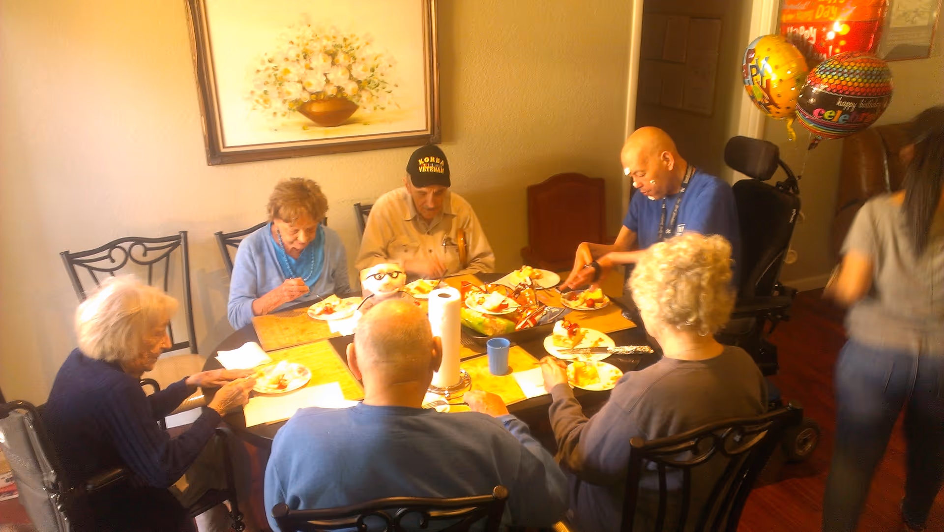 A group of elderly residents sit around a dining table in a senior living facility eating cake with celebration balloons nearby.