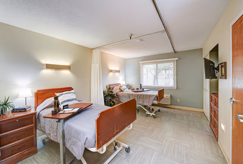 Shared patient bedroom with two adjustable hospital beds, nightstands, a window, and a wall-mounted TV.