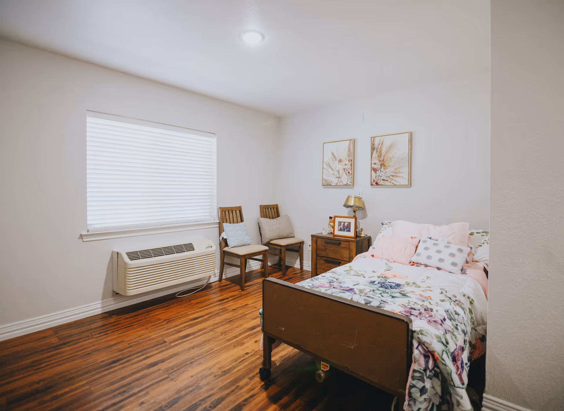 Small bedroom with a single bed covered in floral bedding, a nightstand with a lamp and photos, two chairs, and a window with blinds over a wall-mounted air unit.